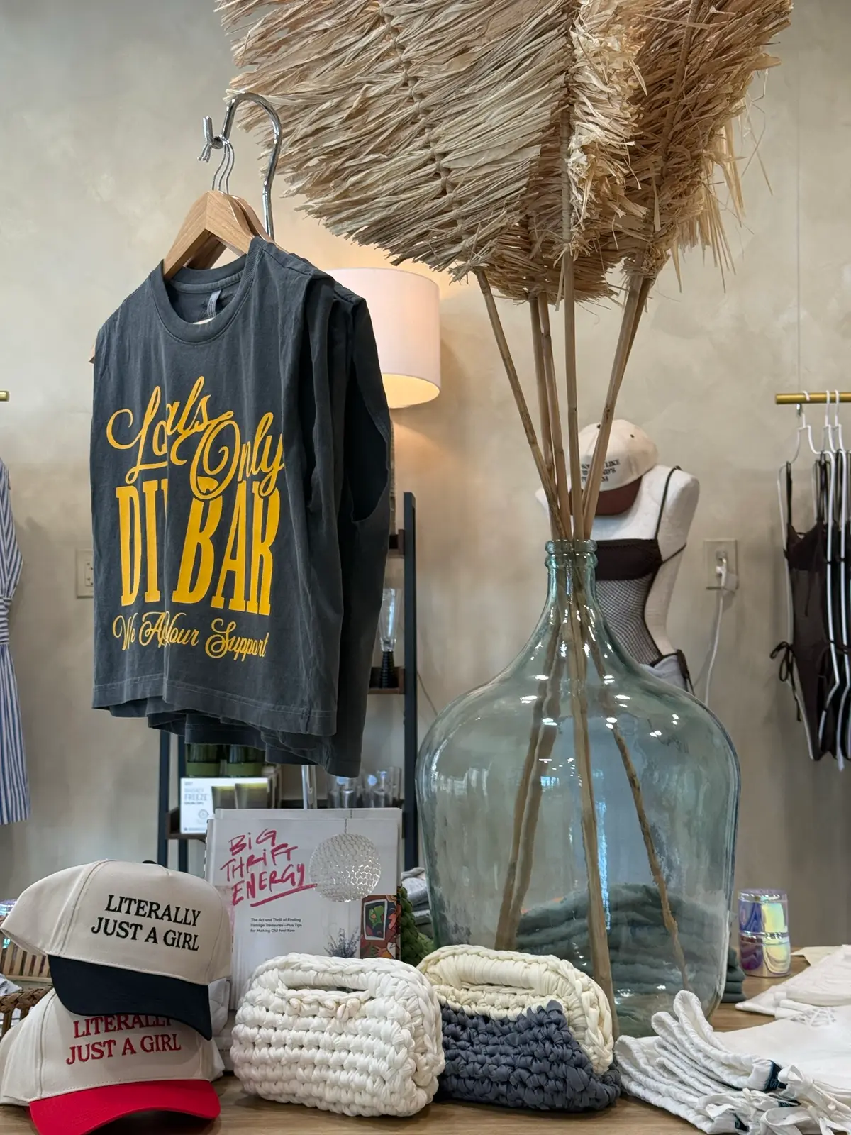 The Trio boutique display table: a graphic tee on a hanger, trucker hats, hand-knit clutches, and dried palms in a glass demijohn