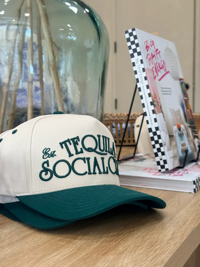 A cream and green Tequila Social Club trucker hat on the boutique table beside stacked books