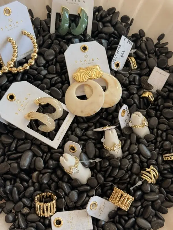 Gold hoops, cuffs, stacking rings, and marbled resin earrings from Sahira Jewelry displayed on black river stones